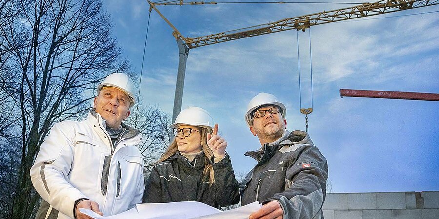 Drei Mitarbeiter auf einer Baustelle mit Baukran im Hintergrund. Sie schauen sich einen Plan an.