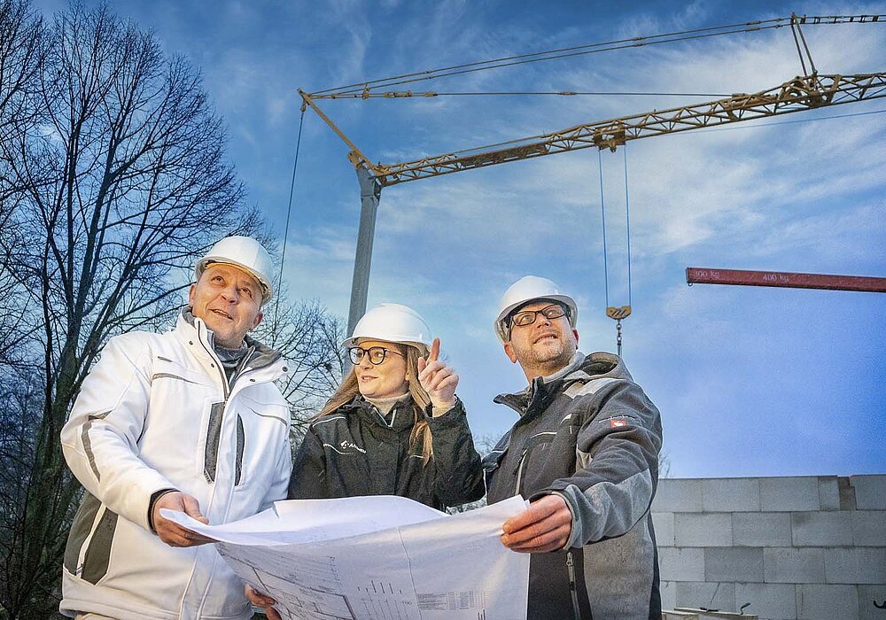 Drei Mitarbeiter auf einer Baustelle mit Baukran im Hintergrund. Sie schauen sich einen Plan an.